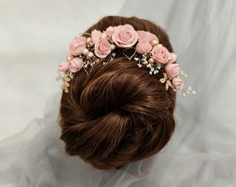 May include: A hair accessory featuring a cluster of small, dusty pink artificial roses interspersed with cream-coloured pearls and delicate white sprigs, arranged in a curved shape to sit atop a brown hair bun.
