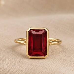 Pode incluir: Um anel de ouro com uma grande pedra preciosa vermelho-escura de corte esmeralda retangular, montada num engaste liso. A banda é fina e polida, ligando-se ao engaste central com um design de haste dividida que se estreita para trás.