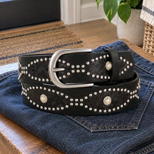 May include: A black leather belt featuring a silver-toned metal buckle, rows of small silver studs arranged in an oval pattern, and circular white faux pearl accents set within silver-toned frames along the length of the strap.