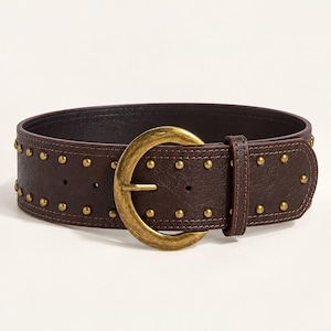 May include: A wide dark brown belt featuring a textured surface, accented with two parallel rows of small round brass-toned metal studs. The belt includes a large, rounded, antique brass-toned metal buckle and a matching leather belt loop.