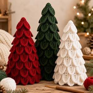 May include: Three cone-shaped decorative trees crafted from yarn using a crochet technique. The trees are arranged in a row, featuring a red tree on the left, a dark green tree in the centre, and a white tree on the right, all with layered petal-like textures.