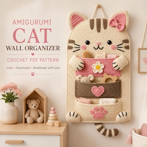 Puede incluir: Un organizador de pared de ganchillo beige con forma de gato y tres bolsillos. El bolsillo superior es rosa con una flor blanca, el central es marrón con un corazón rosa y el inferior es beige con una huella de pata rosa. Texto: AMIGURUMI CAT WALL ORGANIZER.