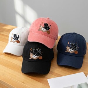 May include: A collection of four baseball caps in white, pink, black, and navy blue. Each cap features an embroidered design of a black cat kneading dough on a wooden board, accompanied by a small pumpkin, a crescent moon, and tiny stars.