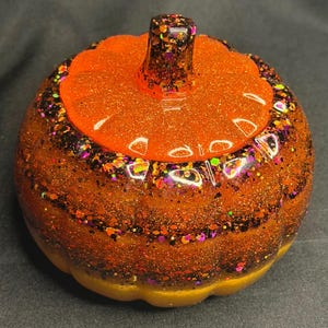 May include: A decorative pumpkin figurine featuring a glossy finish with a vibrant orange top and a base layered with dark orange, black, and purple glitter. The stem is dark with multi-colored glitter accents, and the surface is smooth.