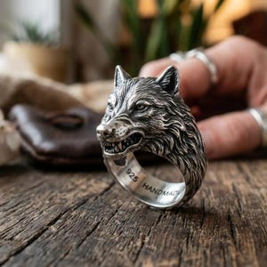 Pode incluir: Um anel de prata com um design detalhado de uma cabeça de lobo a rosnar. A banda tem gravado o texto 925 e HANDMADE na superfície interior. O metal tem um acabamento polido com áreas rebaixadas escurecidas para destacar o pelo e os traços faciais.