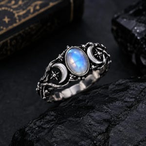 May include: A silver ring featuring a central oval moonstone cabochon flanked by two crescent moon motifs and small star engravings. The band has a textured, vine-like design with oxidized detailing that highlights the intricate metalwork.