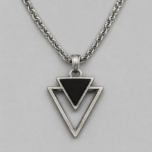 Op de afbeelding: Een zilverkleurige metalen ketting met een hanger in een dubbel driehoekig ontwerp. De binnenste driehoek is gevuld met een effen zwarte inleg, terwijl de buitenste driehoek een open frame is. De hanger hangt aan een dikke, getextureerde ketting.