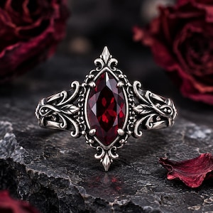 May include: A silver ring featuring a central marquise-cut deep red gemstone. The metal band is crafted with intricate scrollwork and leaf-like filigree details surrounding the stone, creating a pointed, elongated shape on the finger.
