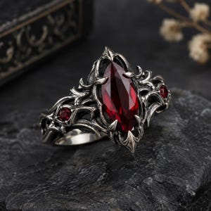 Puede incluir: Un anillo de plata con una gran piedra preciosa de color rojo oscuro en talla marquesa en el centro, flanqueada por dos piedras rojas redondas más pequeñas. La banda metálica tiene un diseño intrincado y texturizado con un acabado oxidado oscuro.