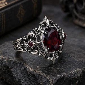 May include: A silver ring featuring an oval-cut deep red gemstone as the centrepiece, surrounded by intricate metal filigree work. Smaller round red stones are set into the ornate, textured silver band, which has a dark, oxidised finish.