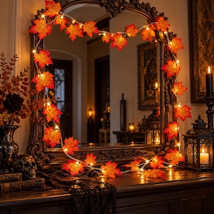 Può includere: Una catena di luci LED con foglie d'acero artificiali arancioni e rosse, drappeggiata attorno alla cornice di un grande specchio. Le luci emettono un bagliore caldo, mettendo in risalto le venature e le texture dettagliate delle foglie.