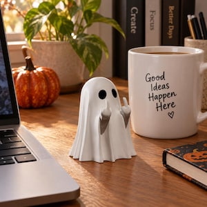Op de afbeelding: Een wit spookfiguurtje met zwarte ovale ogen en twee grijze armen omhoog, elk met een middelvingergebaar. Het spookje staat op een houten oppervlak tussen een witte keramische mok met de tekst Good Ideas Happen Here en een laptop.