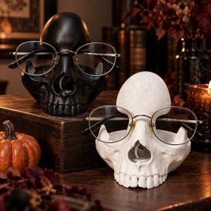 Könnte beinhalten: Zwei brillenförmige Halter in Totenkopfform, einer in Mattschwarz und einer in gesprenkeltem Off-White, jeweils mit einer dünnrandigen Metallbrille, die auf dem Nasenrücken ruht. Die Totenköpfe sind mit realistischen Zähnen und Augenhöhlen geformt.