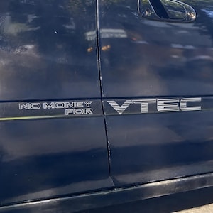 May include: A dark blue car door panel featuring a white vinyl decal that reads NO MONEY FOR VTEC in a stylized font. The text is split across the door seam, with NO MONEY FOR on the left and VTEC on the right, set against the glossy automotive paint.