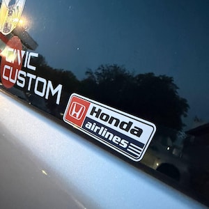 May include: A rectangular Honda Airlines sticker with a red square containing a white H logo, black text, and blue stripes. Next to it is a circular sticker with a cartoon character and a red sticker with the white text CIVIC CUSTOM.