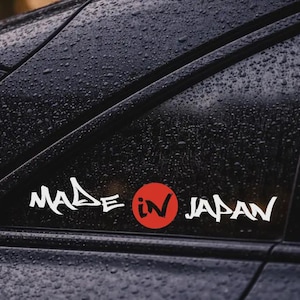Puede incluir: Una pegatina de vinilo blanco aplicada en la ventanilla oscura de un coche, con el texto "MADE IN JAPAN" en una fuente angular estilizada. La palabra "IN" está encerrada en un círculo rojo sólido, imitando el diseño de la bandera japonesa.