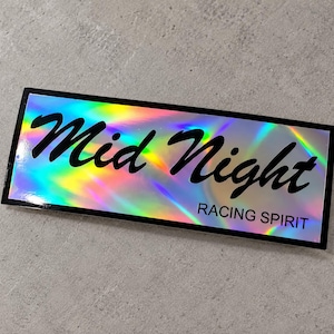 Puede incluir: Una pegatina rectangular con una superficie holográfica que refleja los colores del arcoíris. La pegatina presenta el texto negro Mid Night en una fuente cursiva grande, con las palabras RACING SPIRIT impresas en una fuente más pequeña.