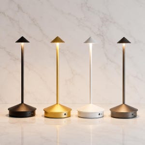 Puede incluir: Cuatro lámparas de mesa portátiles con un tallo delgado y una pantalla cónica. Las lámparas tienen acabados en negro mate, oro, blanco y bronce. Cada lámpara cuenta con una base circular con un puerto de carga visible en el lateral y emite una luz suave.