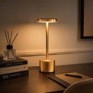 May include: A gold-toned metal table lamp with a cylindrical base, a slender stem, and a flat circular top that emits a warm downward light. The lamp stands next to a stack of three books and a glass reed diffuser with black sticks.