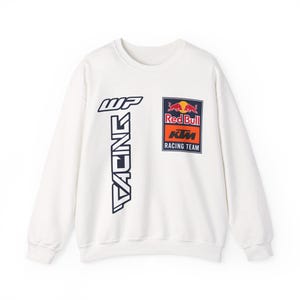 Könnte beinhalten: Ein weißes langärmeliges Sweatshirt mit Rundhalsausschnitt, das auf der linken Seite eine vertikale dunkelblaue Grafik mit dem Text WP RACING und auf der rechten Brustseite einen rechteckigen Red Bull KTM RACING TEAM Aufnäher zeigt.