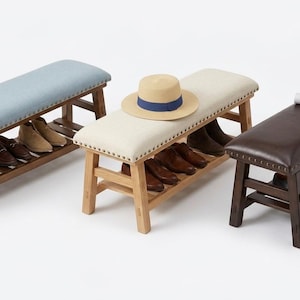 Può includere: Tre panche rettangolari in legno con sedute imbottite e ripiani inferiori per riporre le scarpe. Le panche presentano cuscini color azzurro, crema e marrone scuro con finiture a borchie. Un cappello di paglia è appoggiato sulla panca color crema.