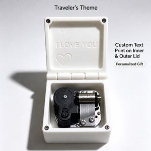Op de afbeelding: Een witte muziekdoos met een open deksel met de in reliëf aangebrachte tekst I LOVE YOU en een hartpictogram. De binnenkant bevat een zilverkleurig en zwart mechanisch muziekwerk. De tekst Outer Wilds Music Box Traveler's Theme is zichtbaar.