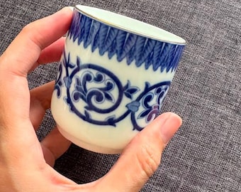 May include: A small ceramic cup with a white base and a blue decorative pattern. The upper rim features a dark blue leaf-like design, while the center displays a blue scrolling vine motif with subtle purple accents. The top edge has a thin metallic trim.