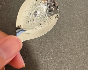 May include: A polished silver-tone metal spoon with a wide, rounded bowl. The surface of the bowl features an intricate, embossed design depicting a central pineapple surrounded by detailed foliage, leaves, and small circular fruit clusters.