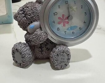 May include: A gray textured teddy bear figurine holding a round silver analog clock. The clock face is light blue with black numbers, a pink flower, and a yellow butterfly. The bear has a blue nose and the text Me to you on its right foot.