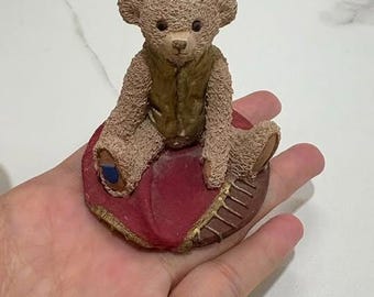 May include: A small, textured beige teddy bear figurine sitting on a circular base. The bear wears a metallic gold vest and is positioned on a dark red, cushion-like surface with a brown, ribbed edge. One paw pad is blue, and the other is brown.