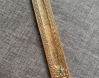 May include: A long, narrow, copper-toned metal incense holder with a rounded top and bottom. The surface features an embossed tree design at the top, a central channel for incense sticks, and intricate floral and vine patterns along the edges.