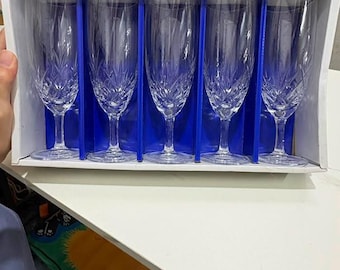 May include: A set of five clear glass champagne flutes arranged in a white cardboard box with blue dividers. Each flute features a tall, slender bowl with a decorative cut-glass pattern at the base and a clear stem leading to a circular foot.