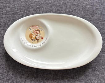 May include: An oval white ceramic dish featuring a circular indentation on the left side. The indentation contains a graphic of two children with the text Ovaltine Biscuits and the phrase The Good Companions printed in black lettering below it.
