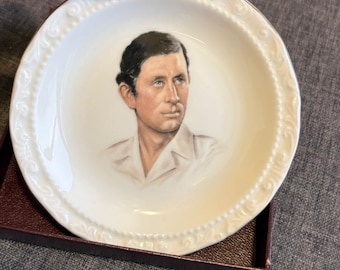 May include: A round white porcelain decorative plate featuring a detailed portrait of a man with dark hair wearing a light-colored collared shirt. The plate has a raised, textured beaded and scrollwork border along the outer edge of the rim.