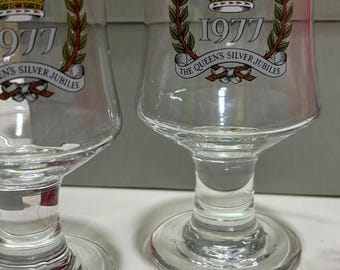 May include: Two clear glass goblets featuring a commemorative design for the 1977 Queen's Silver Jubilee. Each glass displays a colorful crown graphic, green laurel branches, and a banner with the text 1977 THE QUEEN'S SILVER JUBILEE.