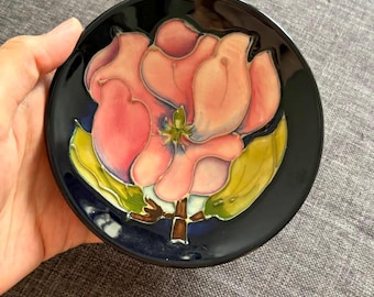 May include: A small, round, black ceramic dish featuring a raised, hand-painted design of a large pink flower with green leaves. The floral motif is centered on the dark background, with visible glossy glaze and defined outlines around the petals.
