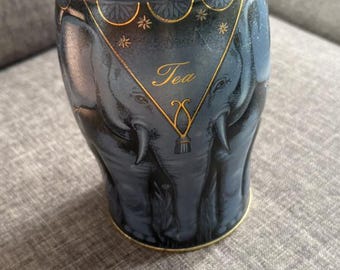 May include: A cylindrical blue metal tea canister featuring a decorative gold elephant illustration and a matching lid. The text "FINE TEAS SINCE 18" is printed on the lid rim, and the word "Tea" is written in gold script on the front.