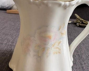 May include: A cream-colored ceramic pitcher featuring a scalloped rim with a gold-tone trim. The body of the pitcher is decorated with a soft, muted floral pattern in shades of pale pink, yellow, and light green, and includes a curved handle.