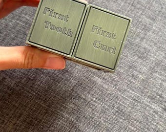 May include: A rectangular, metallic keepsake box with a brushed finish, divided into two separate compartments. The left lid is engraved with the text First Tooth, and the right lid is engraved with the text First Curl, featuring a clean design.