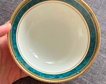 May include: A round ceramic bowl with a smooth white interior and a wide teal marbled rim. The rim features a gold-colored edge and a decorative geometric pattern in black and gold, creating a distinct border around the center of the bowl.
