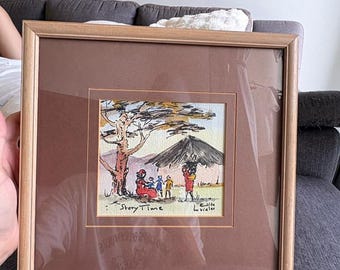 May include: A framed watercolor painting titled Story Time, featuring a group of figures near a tree and a hut. The artwork is enclosed in a light wood frame with a brown matting. The scene depicts individuals in colorful clothing outdoors.