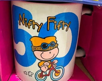 May include: A white ceramic mug featuring a large blue number 50 and the text Nifty Fifty in black. A cartoon character wearing an orange hat and blue goggles rides a red bicycle. The mug is housed in a blue and pink cardboard display box.