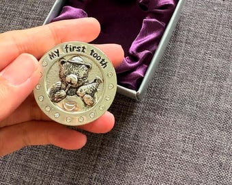May include: A round silver-toned metal keepsake box featuring a relief of a teddy bear and the text "My first tooth" on the lid, accented with small clear crystals along the border, presented in a gray box with a purple satin lining.