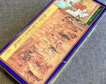 May include: A rectangular blue metal tin featuring a historical illustration of John Cadbury's Shop on Bull Street in Birmingham in the 1830s. The lid includes the text Cadbury's Milk Chocolate Biscuits and depicts people in period clothing.