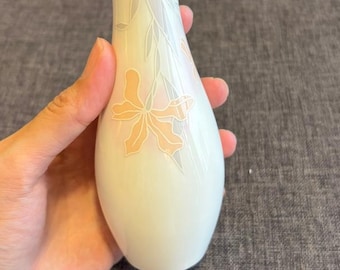 May include: A slender, off-white ceramic bud vase with a narrow neck and a flared, scalloped rim. The surface features a delicate, muted floral pattern in soft peach and pale grey tones, depicting stylized leaves and a single blossom.