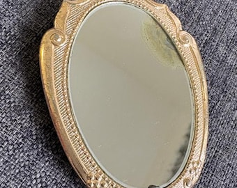 May include: A handheld oval mirror with a metallic gold-tone frame featuring intricate embossed floral and leaf patterns. The mirror surface is reflective with a visible area of discoloration near the top right edge of the glass pane.