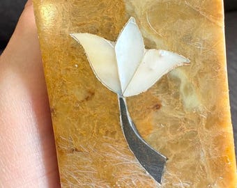 May include: A rectangular, polished amber-colored stone block featuring an inlaid white shell flower design with three petals and a dark, curved stem. The stone has a smooth, glossy surface with natural veining and a translucent, warm golden appearance.