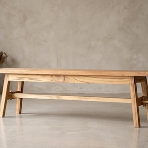 Könnte beinhalten: Eine rechteckige Holzbank mit einer natürlichen, hellbraunen Holzmaserung. Die Bank verfügt über vier angewinkelte Beine, die auf jeder Seite durch eine horizontale Stützstrebe verbunden sind, mit sichtbaren Verbindungsdetails am Sitzrahmen.