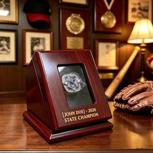 Op de afbeelding: Een donkerhouten vitrine met een schuin voorvenster waarin een kampioensring zit. De basis is voorzien van een in goud gegraveerde tekst: [JOHN DOE] • 2026 STATE CHAMPION. De behuizing heeft een gepolijste afwerking en een kijkvenster.