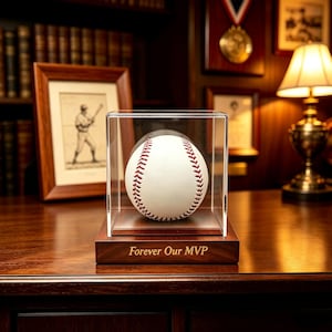 Könnte beinhalten: Ein weißer Baseball mit roten Nähten in einer durchsichtigen Acryl-Vitrine mit einem dunklen Holzsockel. Auf dem Sockel befindet sich die eingravierte Aufschrift Forever Our MVP in goldener Schrift, die als Schutz und Präsentation dient.